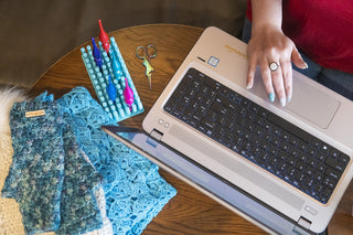 Hand typing on laptop with crocheted pieces and accessories viewed from above.