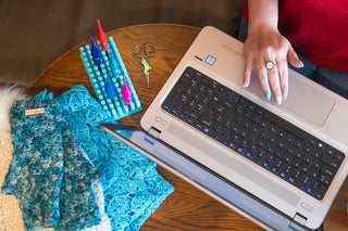Laptop, hand, hooks and crochet accessories on a table, viewed from above.