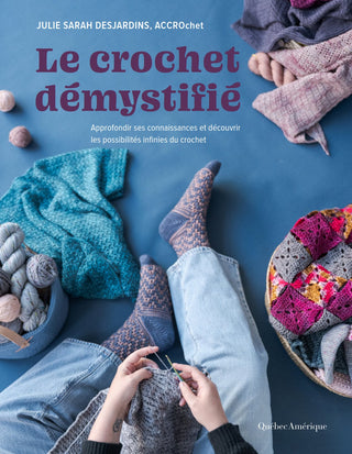French-language crochet book cover titled "Le crochet démystifié" by Julie Sarah Desjardins, Accrochet, featuring a top-down scene of hands crocheting with patterned socks, blue jeans, yarn balls, and knitted textiles on a blue surface.