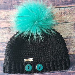 The black crochet hat with teal pompom and buttons on dark background.