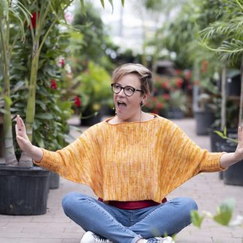 Woman sitting cross legged wearing orange crochet top with large plants behind her