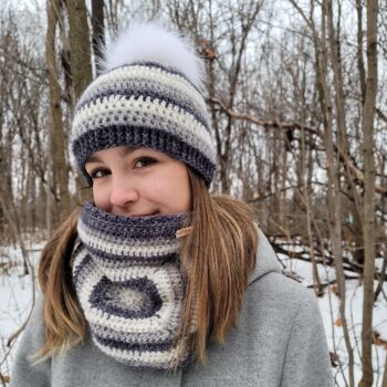 Young woman wearing striped crochet hat with white pompom and cowl with large granny square in the woods